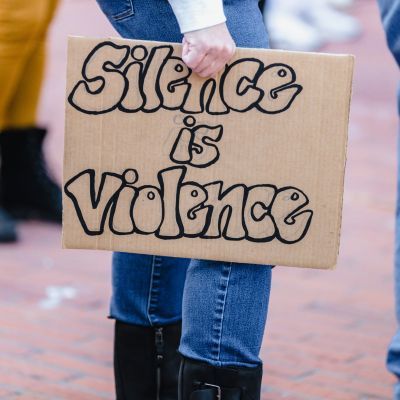Someone holding a cardboard sign that spells out 'Silence is violence' by their legs